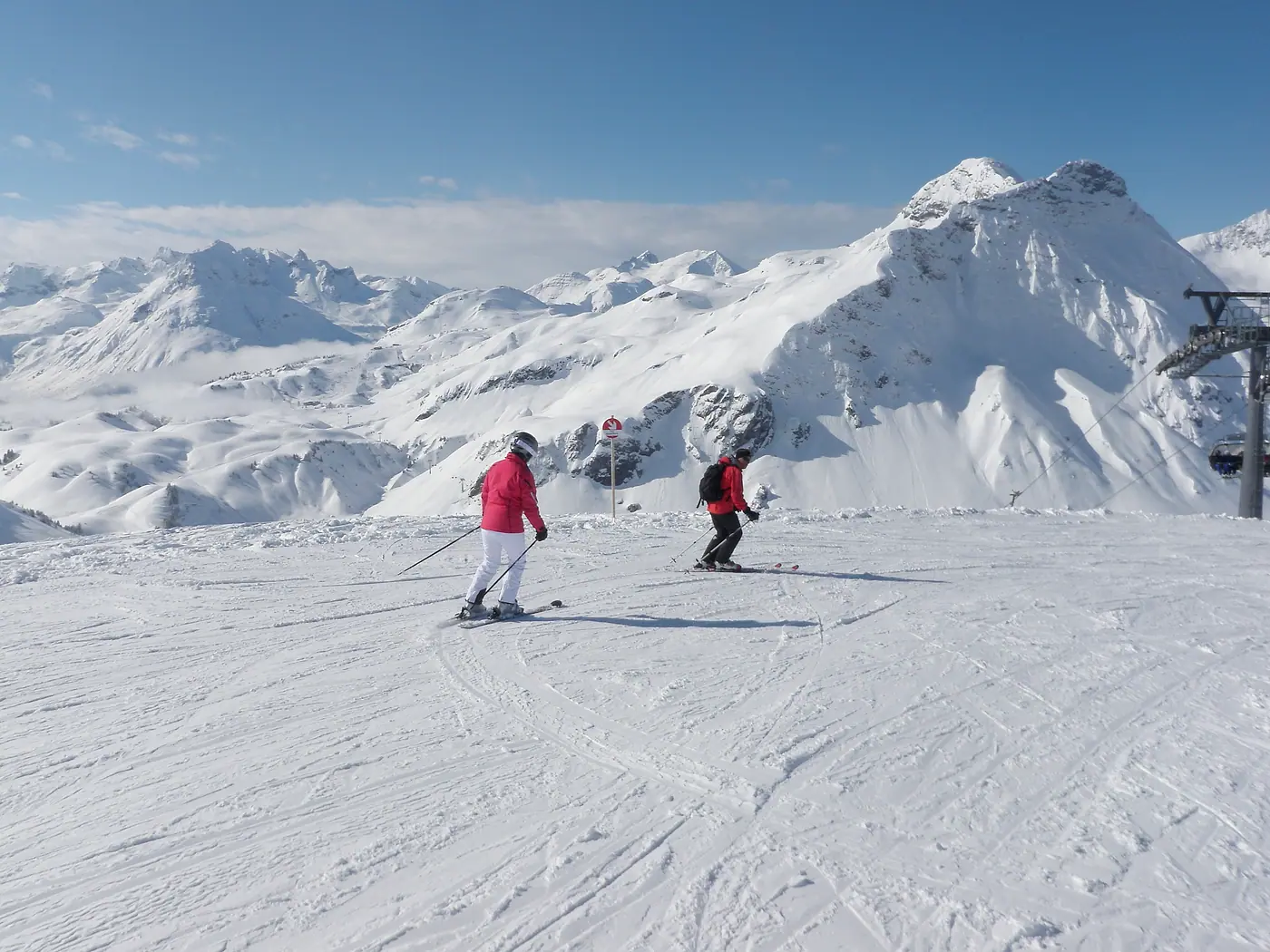 Skilehrer in roter Jacke und ein Gast auf der Piste, dahinter verschneite Gipfel am Arlberg