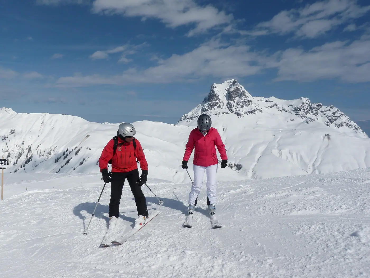 Skilehrer in roter Jacke und eine Skifahrerin auf der Piste, dahinter ein Gipfel im Schnee